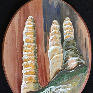 May include: Oval wooden wall art featuring a painted cave scene with three stalagmites in shades of beige, tan, and green. The background has vertical stripes of brown, blue, and gray. The artwork is signed in the lower right corner.