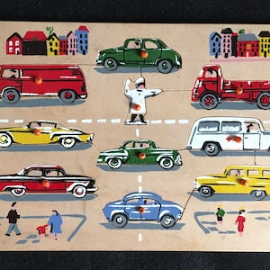 May include: A wooden toy puzzle featuring a traffic scene with a variety of cars, trucks, and a traffic officer. The puzzle pieces are painted in bright colors, including red, yellow, blue, green, and white. The puzzle is set against a background of houses.