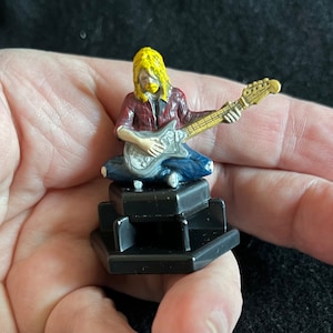 May include: A small plastic figurine of a musician playing a guitar. The figurine is painted in a realistic style with a red and black plaid shirt, blue jeans, and yellow hair. The figurine is standing on a black hexagonal base.