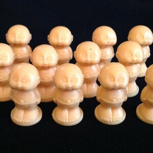 May include: A group of 12 small, peach-colored figurines shaped like babies. Each figurine has a round head, short limbs, and a simple, smiling face.
