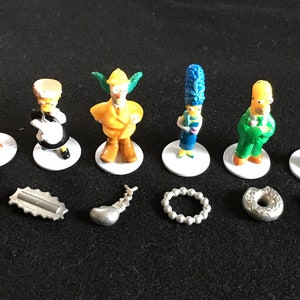 May include: A set of seven Simpsons character figurines, including Bart, Lisa, Marge, Homer, Krusty the Clown, and two others. The figurines are on white bases and come with seven silver accessories, including a saxophone, a donut, a boxing glove, a necklace, a wrench, a pair of scissors, and a metal bar.