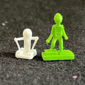 May include: Two plastic game pieces, one white and one green. The white piece is a small figure with arms outstretched, sitting on a small base. The green piece is a standing figure with a helmet, wearing a jumpsuit and standing on a green base.