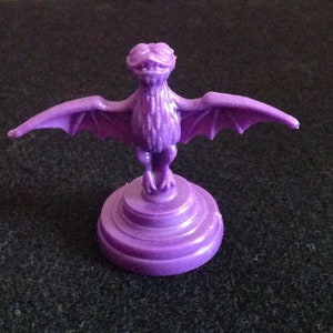 May include: Purple plastic game piece shaped like a bat with outstretched wings standing on a three-tiered base.