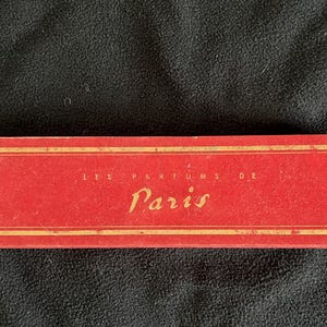 May include: A rectangular red box with gold-colored trim and lettering. The text on the box reads LES PARFUMS DE Paris in a combination of block and script fonts. The box has a flat, elongated shape and a smooth, matte finish on its surface.