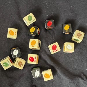May include: A collection of 14 dice with various symbols and colors, including black, white, red, yellow, and green. The dice are on a dark gray fabric surface.