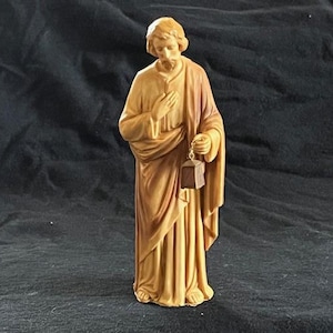 May include: A wooden figurine of a man in a robe, holding a small box in his right hand. The figurine is painted in shades of brown and beige.