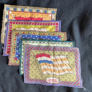 May include: A set of seven vintage fabric patches featuring different flags. The patches are rectangular and have a colorful, patterned border. The bottom patch features a flag with orange, white, and blue stripes and the text "ORANGE FREE STATE".