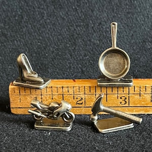 May include: Four miniature silver-colored game pieces: a high-heeled shoe, a motorcycle, a hammer, and a frying pan. The pieces are arranged on a wooden ruler with inch markings.