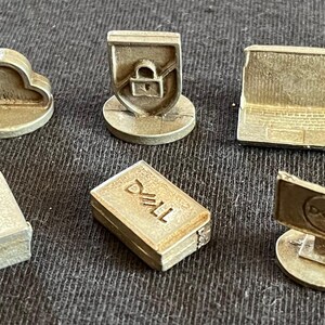 May include: Six small, silver-colored, 3D printed figurines representing different aspects of technology. The figurines include a cloud, a shield with a lock, a laptop, a box with the word "DELL" on it, a desktop computer monitor with the DELL logo, and a box with a textured surface.