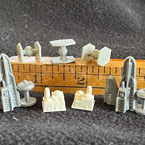 May include: A collection of eight miniature Star Wars spaceship models, including TIE fighters, X-wings, and a Death Star. The models are made of gray plastic and are arranged on a wooden ruler.