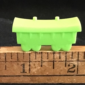 May include: A small, bright green toy train car with a curved top and two small pegs. The car is approximately 1 inch long and has four rounded wheels. It is placed on a wooden ruler with black markings against a dark background.
