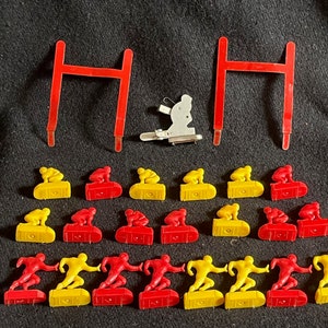 May include: A set of 20 miniature football players, 10 in yellow and 10 in red, with two red goalposts. The players are in a variety of poses, including running, blocking, and kicking. The goalposts are made of metal and have a simple design.