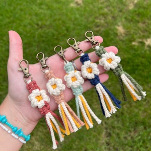 May include: Six colorful crocheted daisy keychains with gold key rings. Each keychain has a white daisy with a yellow center and a tassel made of the same color yarn as the daisy.