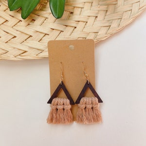 May include: A pair of wooden triangle earrings with a brown wood finish and a light brown tassel. The earrings are hanging from a brown card.