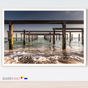 May include: A weathered wooden pier extends into the ocean, with the water lapping at the base of the pilings. The sky is a muted blue, and the water is a calm turquoise.
