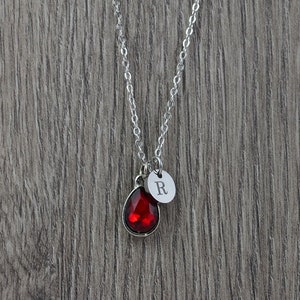 May include: A silver chain necklace with a red teardrop-shaped crystal and a silver charm with the letter "R" on it.