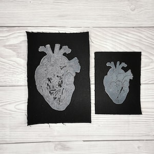 Könnte beinhalten: Zwei schwarze Stoffpatches mit weißen anatomischen Herzdesigns. Der größere Patch hat ein detailliertes Herzdesign, während der kleinere Patch ein einfacheres Herzdesign hat.