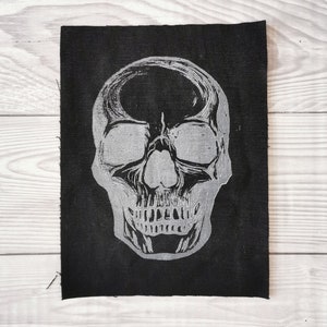 Peut inclure: Un patch en tissu noir avec un motif de crâne argenté. Le crâne est détaillé avec des orbites, un nez et des dents.