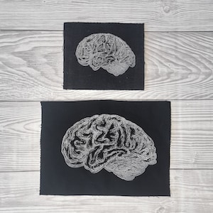 Könnte beinhalten: Zwei schwarze Stoffpatches mit einem weißen gestickten Bild eines menschlichen Gehirns. Die Patches sind unterschiedlich groß.