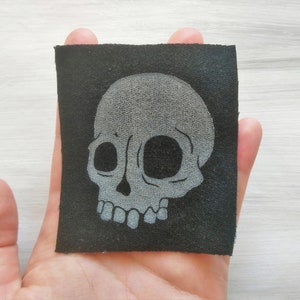 Könnte beinhalten: Ein grauer Totenkopf-Aufnäher auf einem schwarzen Stoff. Der Totenkopf ist weiß umrandet und hat einen einfachen, cartoonartigen Stil.