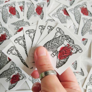 May include: A sheet of temporary tattoos featuring a black and white anatomical heart with a red heart inside. The tattoos are on a white background and are arranged in a stack.