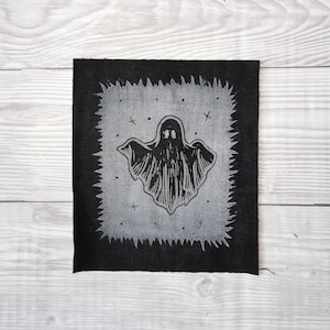 Op de afbeelding: Zwarte stoffen patch met een witte omtrek van een geest met een gekartelde rand. De geest bevindt zich in een wit veld met kleine zwarte sterren en kruisen.