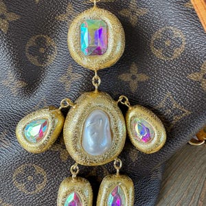 Puede incluir: Un collar con colgante dorado con una gran gema rectangular iridiscente en la parte superior, una piedra central nacarada y varias gemas iridiscentes más pequeñas. El colgante cuelga de una cadena dorada.