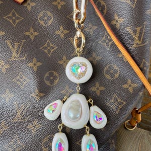 Puede incluir: Un charm blanco y dorado con detalles iridiscentes. El charm presenta una serie de elementos blancos en forma de lágrima conectados por cadenas doradas. El charm está sujeto a un cierre dorado, sobre un fondo marrón.