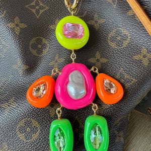 Puede incluir: Un llavero colorido con un cierre dorado. Presenta un círculo verde neón con una gema rosa, una forma irregular rosa con una perla plateada, dos gotas naranjas con gemas y dos rectángulos verdes con gemas. El fondo es marrón.