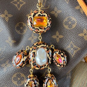 Puede incluir: Un dije dorado con un diseño de estampado de leopardo. El dije presenta una gema grande de color naranja y varias gemas más pequeñas en forma de lágrima. Una perla blanca grande está en el centro del dije.