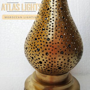 May include: A gold metal Moroccan lantern with a round, perforated body and a wide base. The lantern has a traditional design with intricate cutouts.