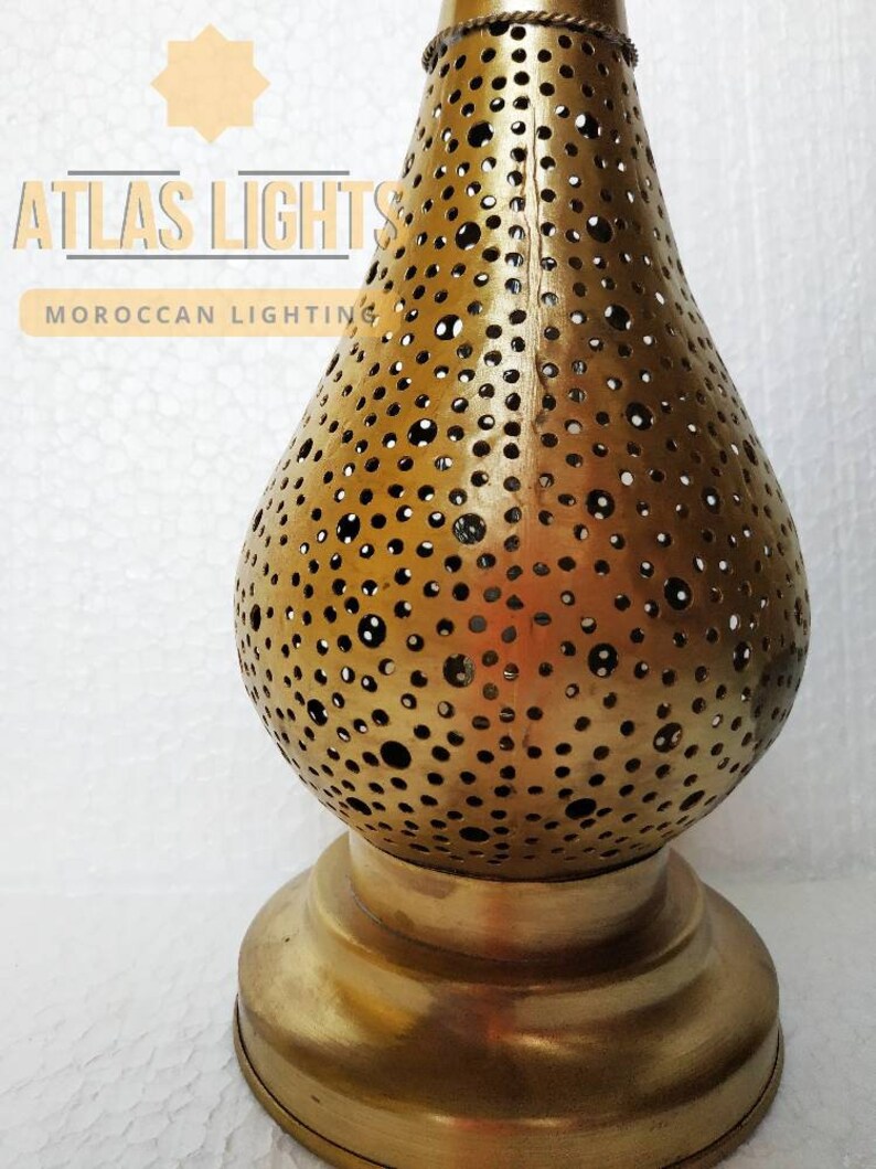 May include: A gold metal Moroccan lantern with a round, perforated body and a wide base. The lantern has a traditional design with intricate cutouts.