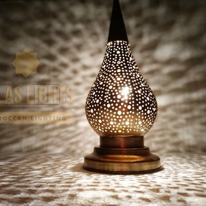 May include: A gold Moroccan table lamp with a pear-shaped shade featuring a delicate, intricate pattern of small holes. The lamp is lit, casting a warm glow through the holes.