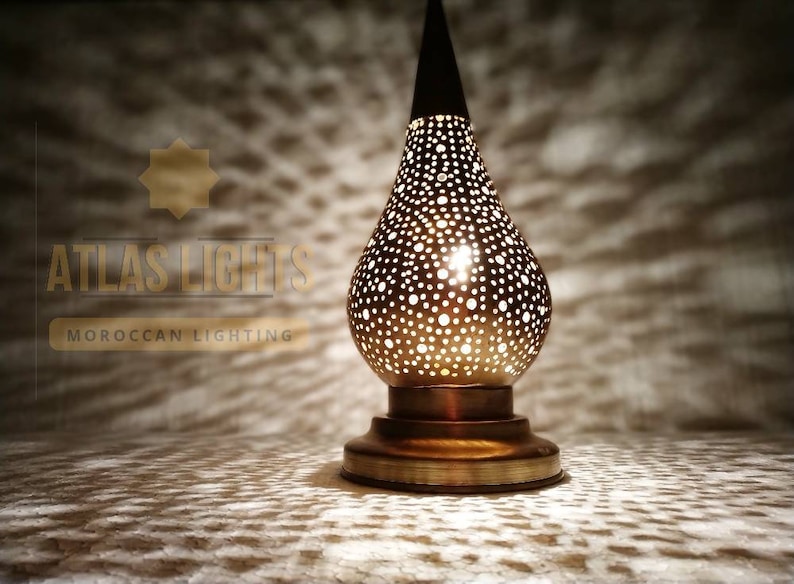 May include: A gold Moroccan table lamp with a pear-shaped shade featuring a delicate, intricate pattern of small holes. The lamp is lit, casting a warm glow through the holes.
