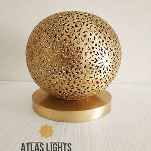 K&ouml;nnte beinhalten: Eine goldene marokkanische Pendelleuchte mit einem runden, filigranen, ausgeschnittenen Design. Die Leuchte steht auf einem runden, goldenen Sockel.