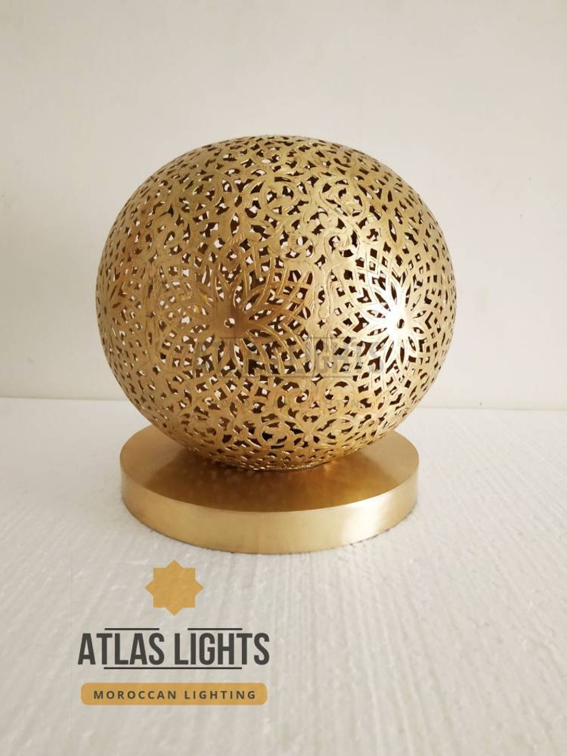 K&ouml;nnte beinhalten: Eine goldene marokkanische Pendelleuchte mit einem runden, filigranen, ausgeschnittenen Design. Die Leuchte steht auf einem runden, goldenen Sockel.