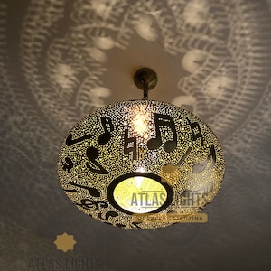 May include: A gold-toned metal pendant light fixture with a round, perforated shade. The shade features a pattern of musical notes and stars. The light is hanging from a ceiling and casting a shadow on the wall.