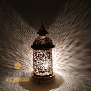 Op de afbeelding: Een gouden Marokkaanse lantaarn met ingewikkelde uitsnijdingen en een ronde basis. De lantaarn is verlicht en werpt een warme gloed op de omgeving. De tekst "ATLAS LIGHTS MOROCCAN LIGHTING" is zichtbaar in de linkerbenedenhoek van de afbeelding.