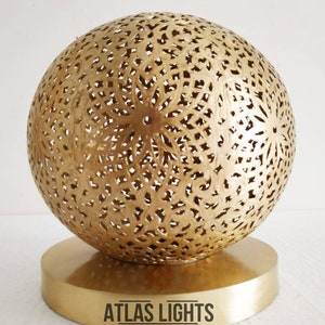 K&ouml;nnte beinhalten: Eine goldene Tischlampe im marokkanischen Stil mit einem runden, filigran gemusterten Schirm und einem runden Sockel. Die Lampe ist aus Metall und hat ein traditionelles Design. Der Text "ATLAS LIGHTS" und "MOROCCAN LIGHTING" ist unterhalb der Lampe sichtbar.