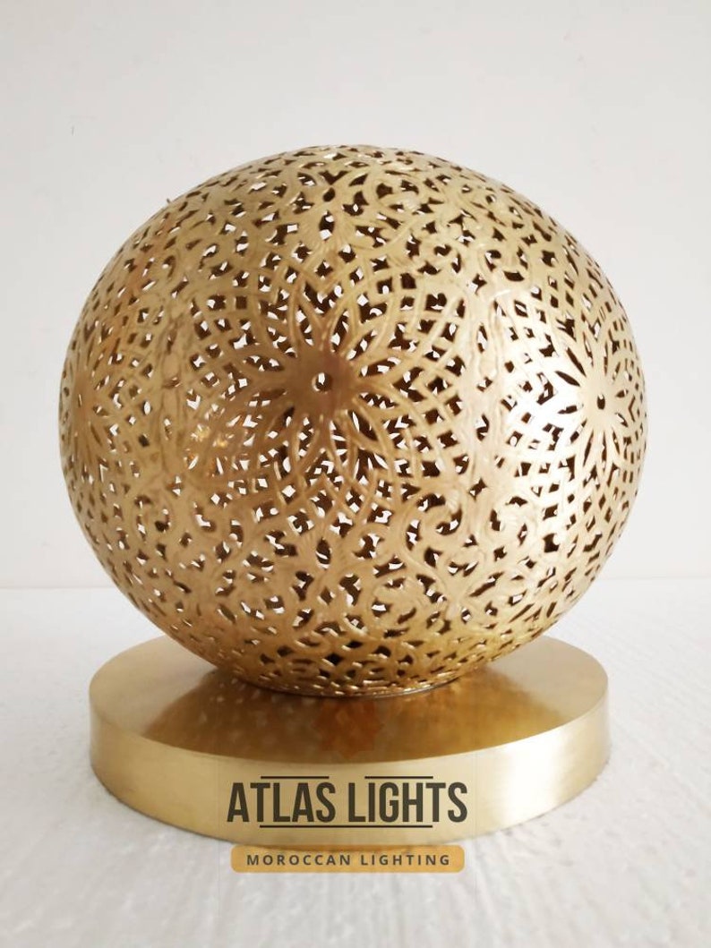 K&ouml;nnte beinhalten: Eine goldene Tischlampe im marokkanischen Stil mit einem runden, filigran gemusterten Schirm und einem runden Sockel. Die Lampe ist aus Metall und hat ein traditionelles Design. Der Text "ATLAS LIGHTS" und "MOROCCAN LIGHTING" ist unterhalb der Lampe sichtbar.