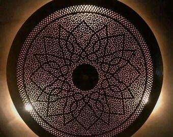May include: A round, decorative ceiling light fixture with a intricate, geometric pattern cut out of the metal. The light is on and casting a warm glow.