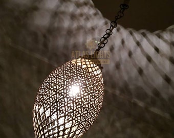Op de afbeelding: Een ovale hanglamp met een gedetailleerde, opengewerkte metalen kap. De lamp hangt aan een ketting en werpt ingewikkelde schaduwen. Het warme licht schijnt door het open ontwerp. De tekst "ATLAS LIGHTS" is zichtbaar.
