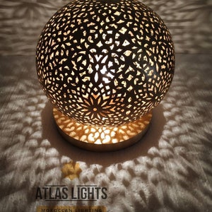 K&ouml;nnte beinhalten: Eine goldene marokkanische Tischlampe mit einem runden, filigranen, ausgeschnittenen Design. Die Lampe ist beleuchtet und wirft Schatten auf die wei&szlig;e Oberfl&auml;che darunter. Der Text "ATLAS LIGHTS MOROCCAN LIGHTING" ist unterhalb der Lampe sichtbar.