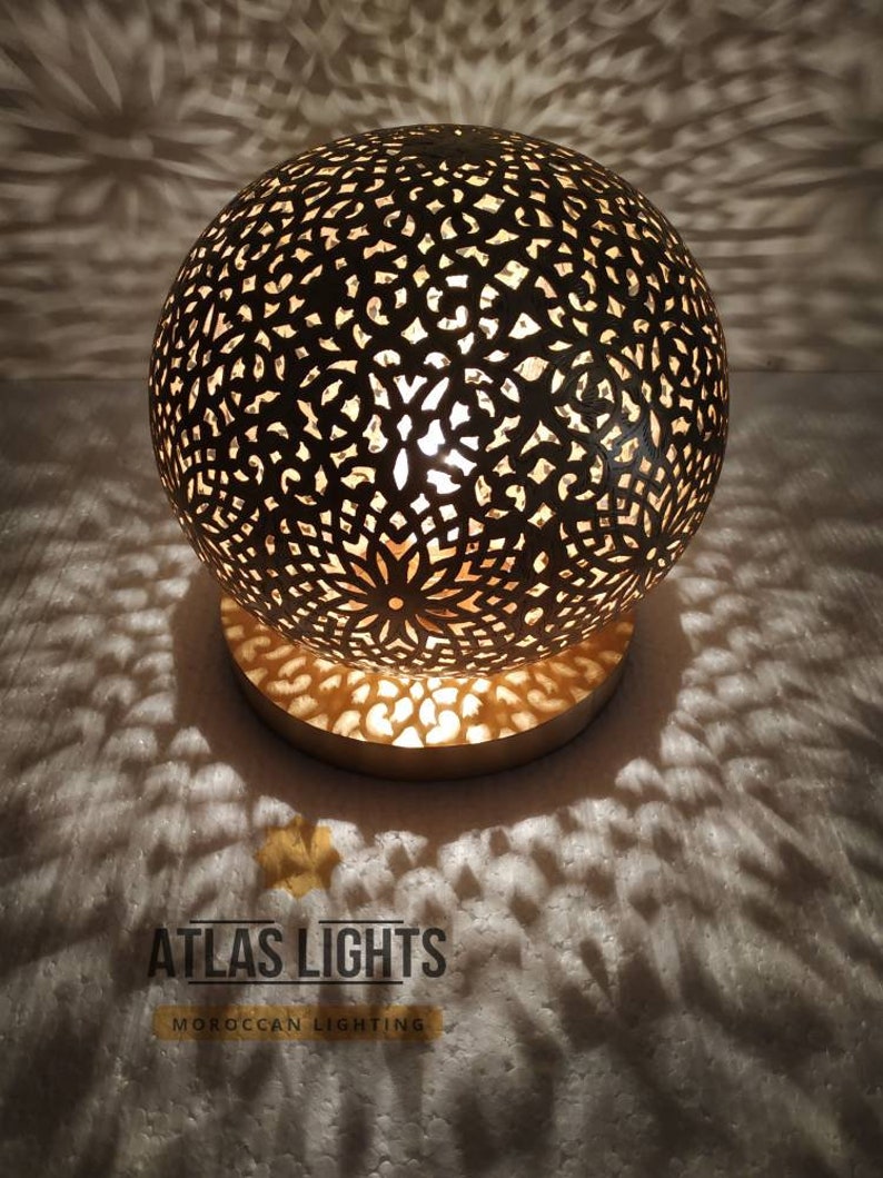 K&ouml;nnte beinhalten: Eine goldene marokkanische Tischlampe mit einem runden, filigranen, ausgeschnittenen Design. Die Lampe ist beleuchtet und wirft Schatten auf die wei&szlig;e Oberfl&auml;che darunter. Der Text "ATLAS LIGHTS MOROCCAN LIGHTING" ist unterhalb der Lampe sichtbar.