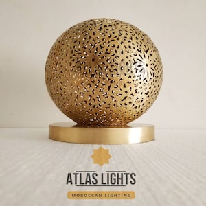 K&ouml;nnte beinhalten: Eine goldene Tischlampe im marokkanischen Stil mit einem runden, filigranen, ausgeschnittenen Design. Die Lampe steht auf einem runden Sockel und tr&auml;gt den Text "ATLAS LIGHTS MOROCCAN LIGHTING" darunter.