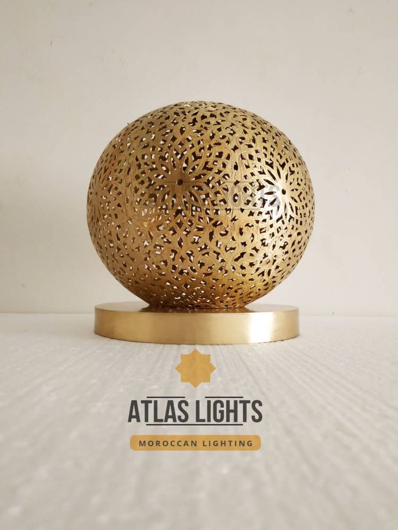 K&ouml;nnte beinhalten: Eine goldene Tischlampe im marokkanischen Stil mit einem runden, filigranen, ausgeschnittenen Design. Die Lampe steht auf einem runden Sockel und tr&auml;gt den Text "ATLAS LIGHTS MOROCCAN LIGHTING" darunter.
