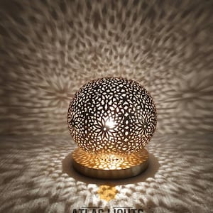 K&ouml;nnte beinhalten: Eine goldene marokkanische Tischlampe mit filigranen Ausschnitten. Die Lampe wirft ein wundersch&ouml;nes Lichtmuster an die Wand hinter sich. Die Lampe steht auf einem runden Sockel und hat einen kugelf&ouml;rmigen Schirm. Die Lampe ist eingeschaltet und das Licht scheint durch die Ausschnitte.