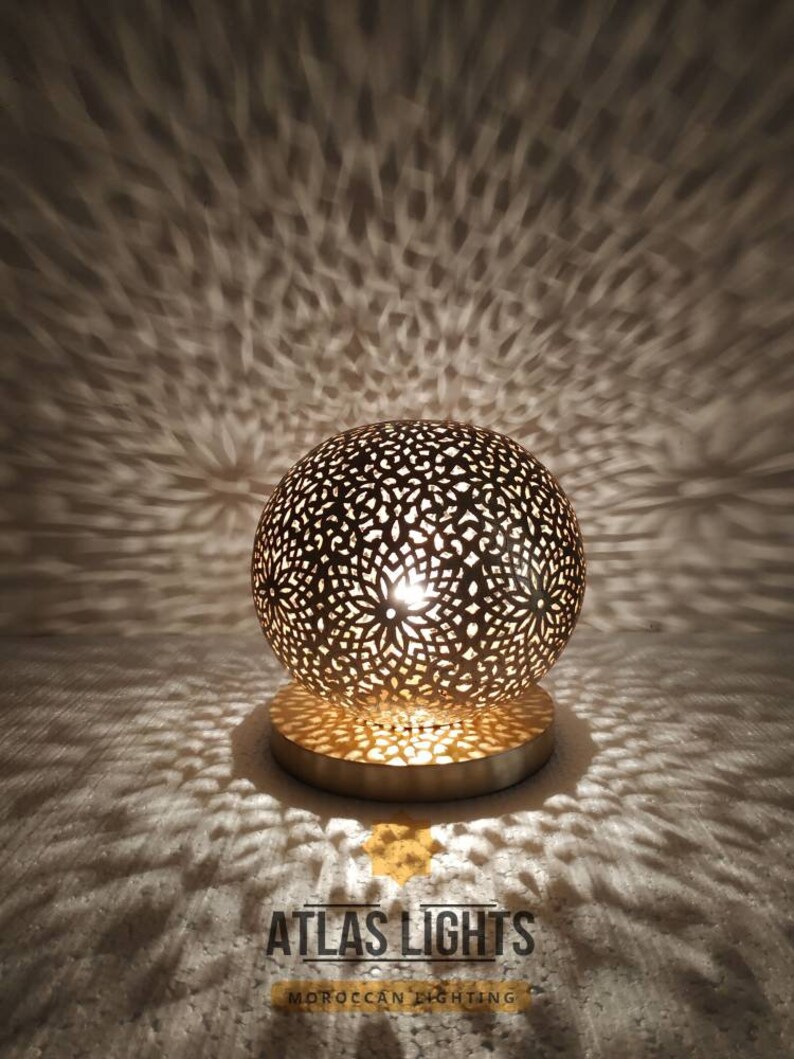 K&ouml;nnte beinhalten: Eine goldene marokkanische Tischlampe mit filigranen Ausschnitten. Die Lampe wirft ein wundersch&ouml;nes Lichtmuster an die Wand hinter sich. Die Lampe steht auf einem runden Sockel und hat einen kugelf&ouml;rmigen Schirm. Die Lampe ist eingeschaltet und das Licht scheint durch die Ausschnitte.