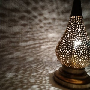 May include: A gold metal lamp with a round, perforated shade. The lamp is lit and casting a pattern of light on the surface below.