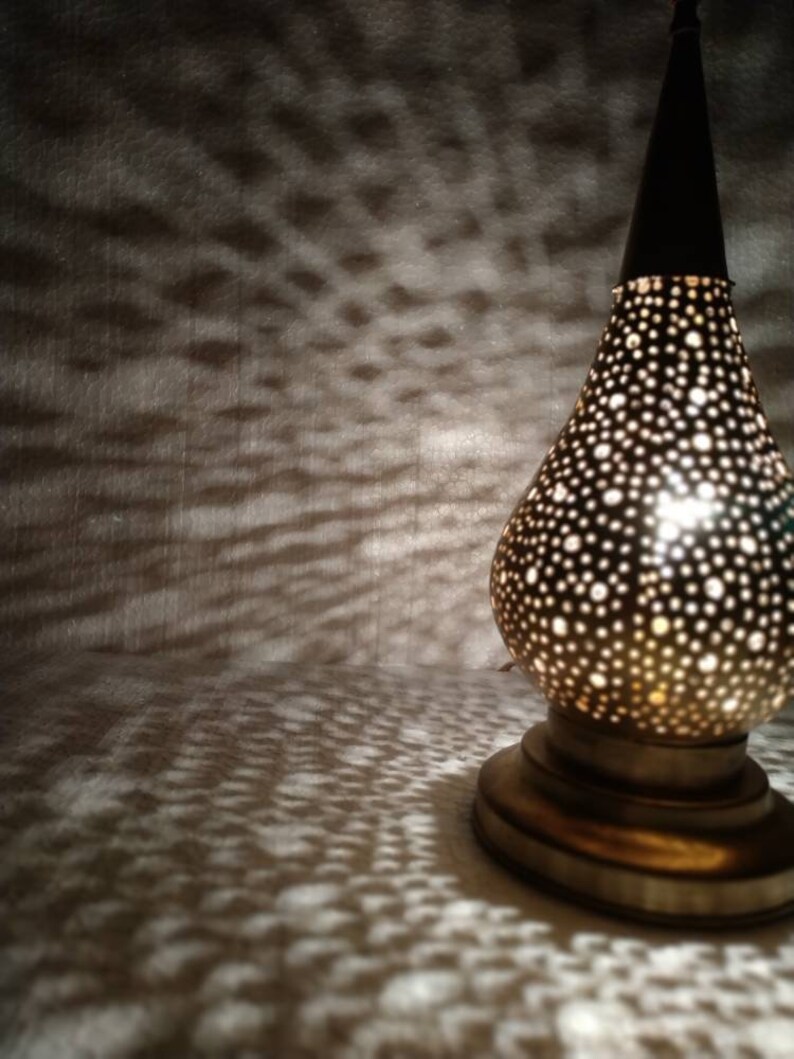 May include: A gold metal lamp with a round, perforated shade. The lamp is lit and casting a pattern of light on the surface below.